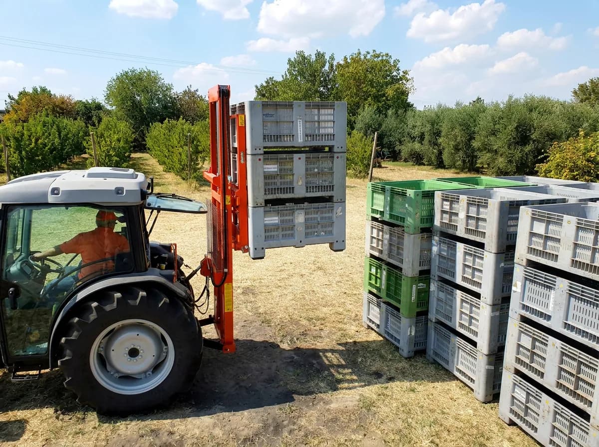 Narrow orchard tractor with a rear-mounted hydraulic fork stacker, lifting a stack of harvest crates beside rows of stacked bins