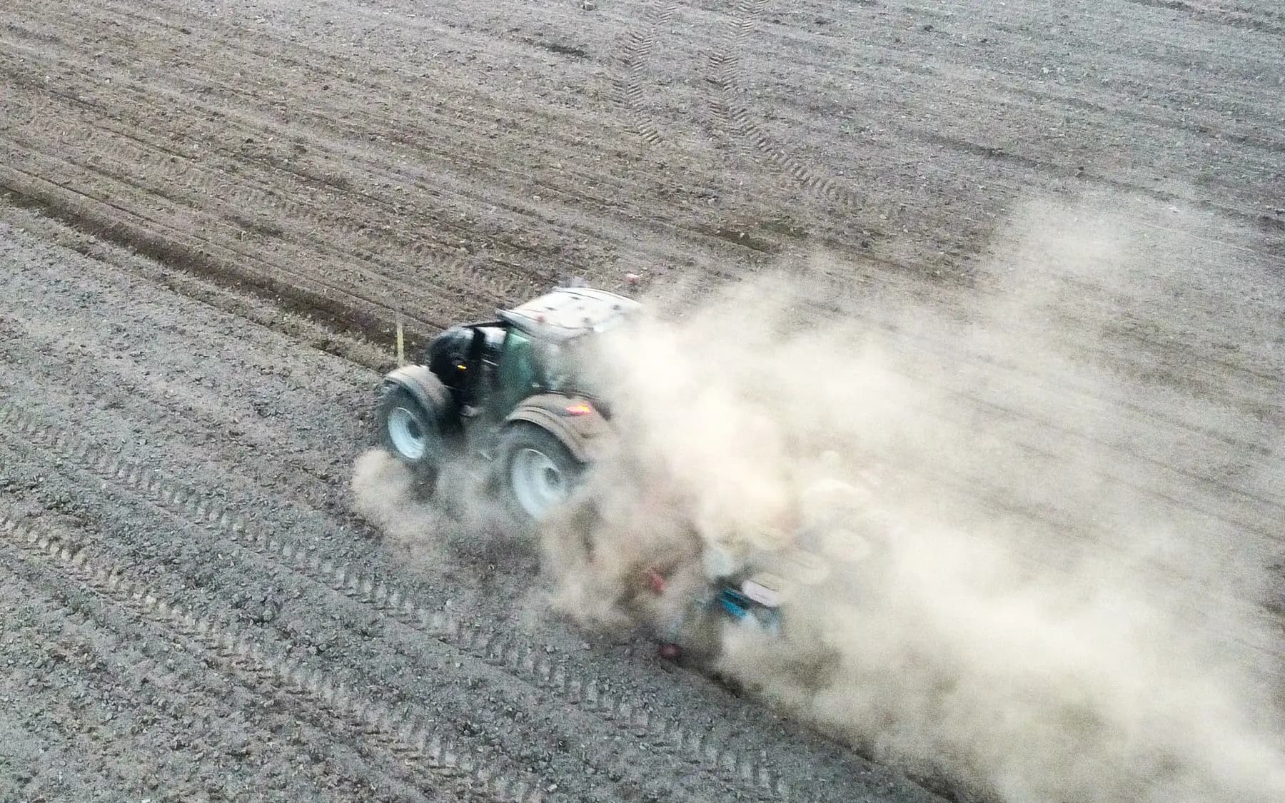 Tractor pulling an implement across a dry field, seen from above, with a cloud of dust behind it.