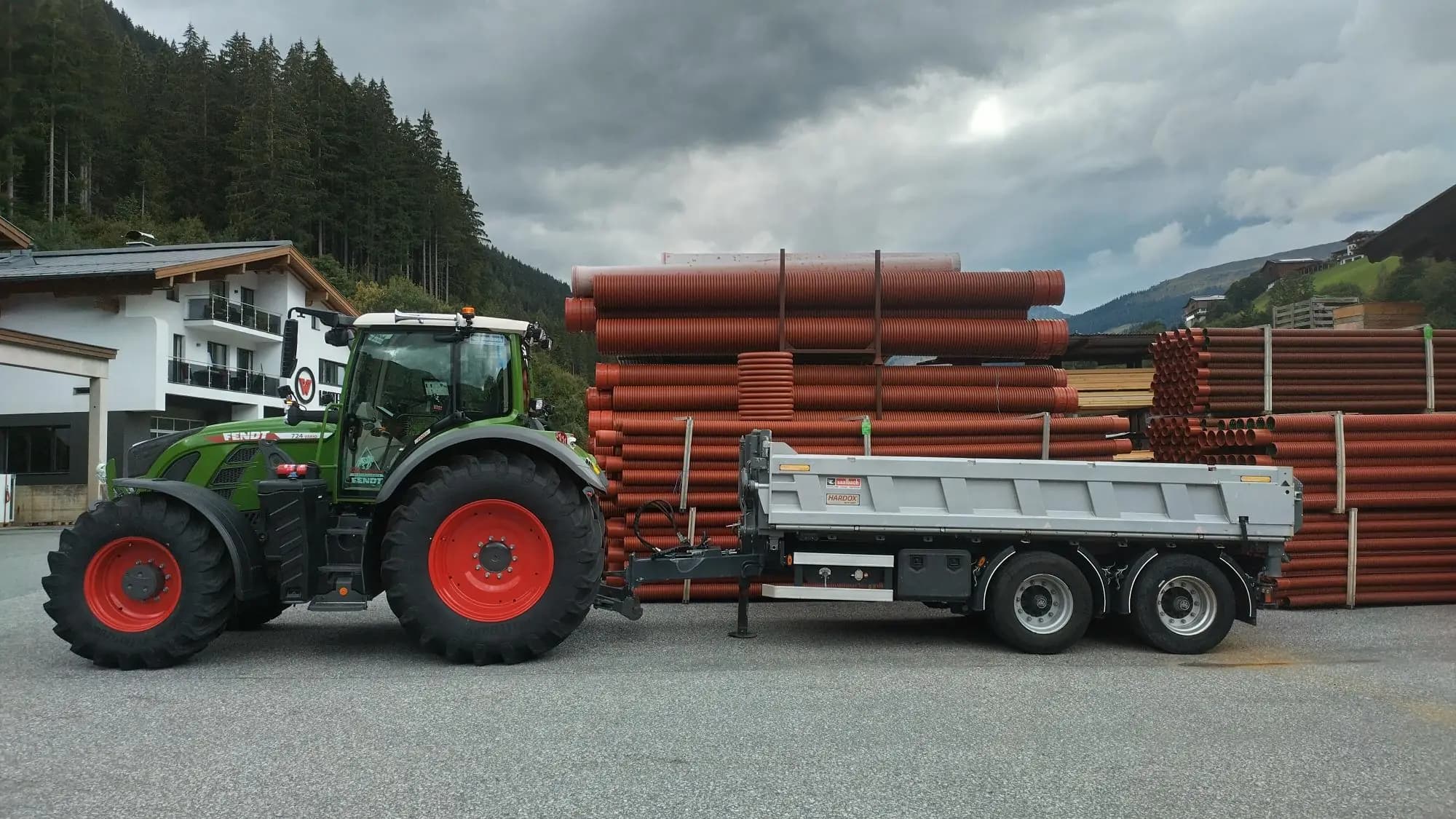 Fendt 724 Vario coupled to a Saalbach three-way tipper trailer in a builder’s yard, orange drainage pipe stacked behind, mountains in the background